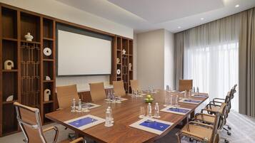 A conference room with a large wooden table, beige chairs, a projector screen, and a large window with curtains.