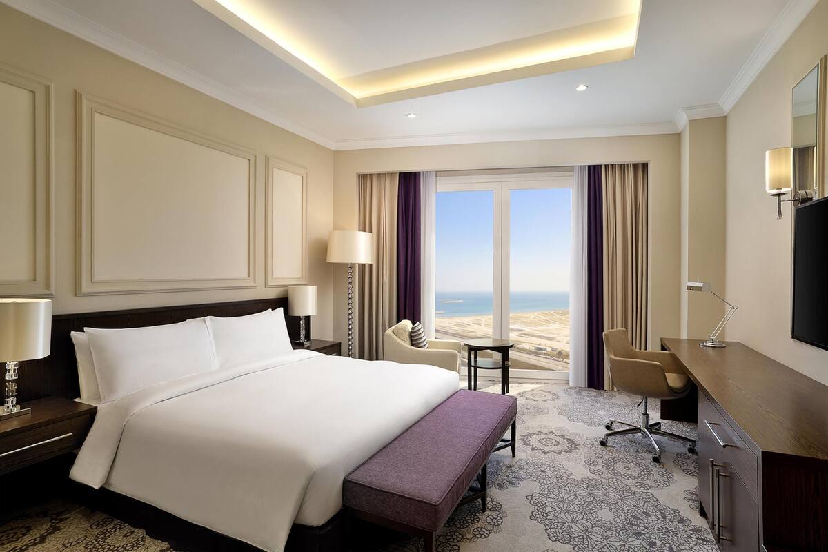 Quarto de hotel com uma cama grande, uma escrivaninha, uma cadeira, um banco, uma televisão e vista para a praia.