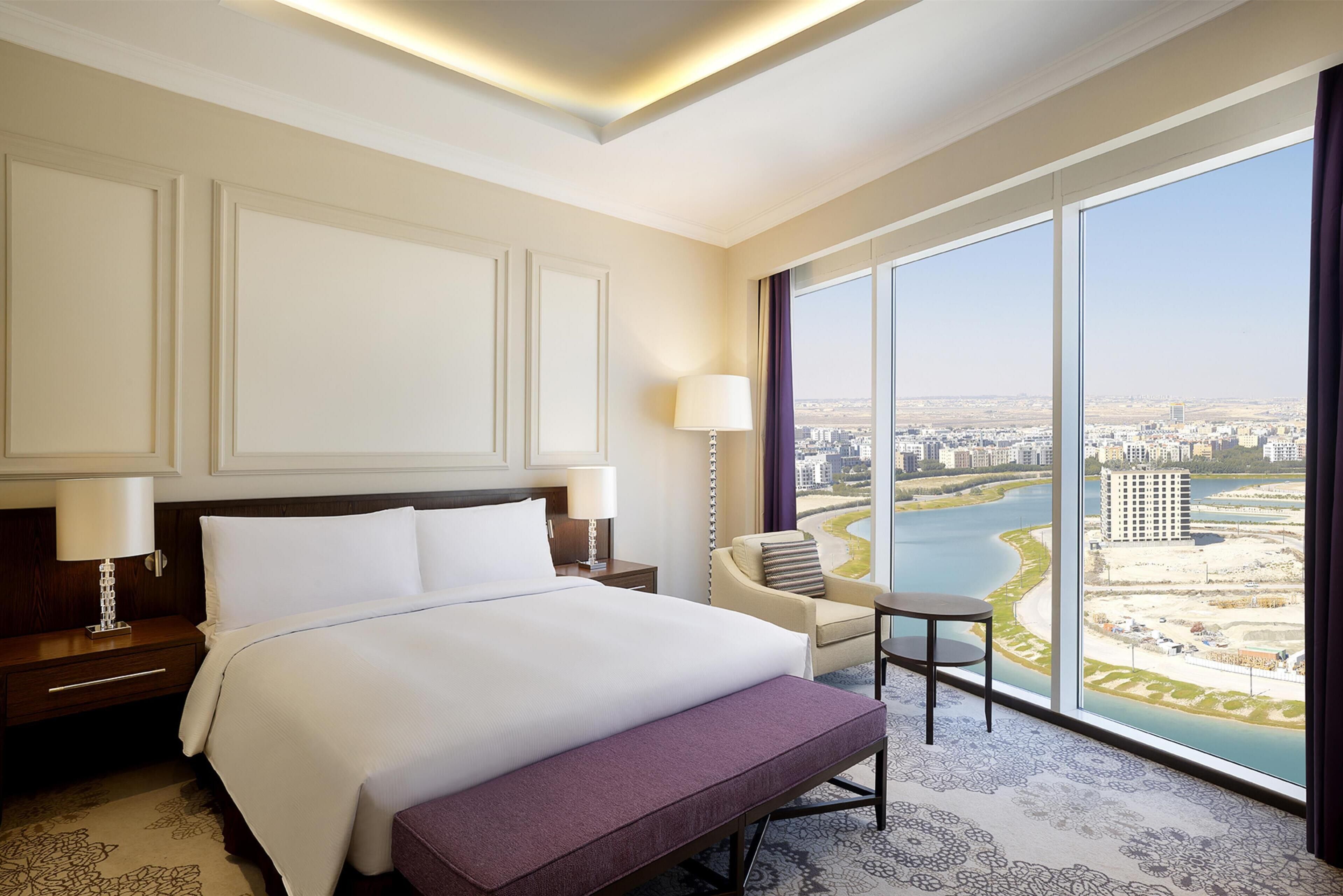 A hotel room with a large bed, a chair, a small table, and a view of a river and cityscape through large windows.