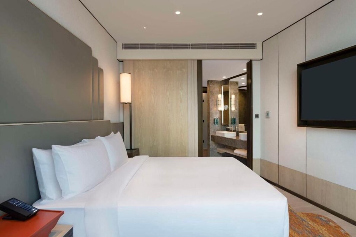 A hotel room with a large bed, a bedside table with a remote control, a wall-mounted TV, and a bathroom visible through an open door.