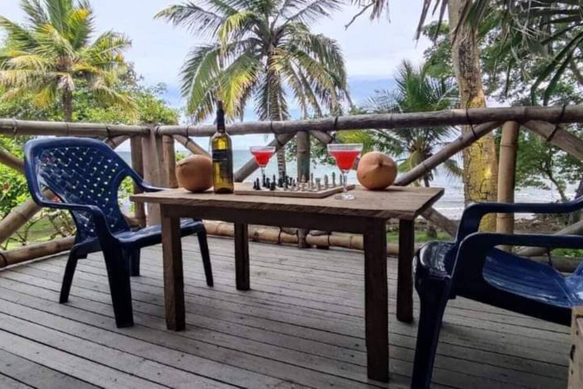 Una terraza de madera con dos sillas de plástico azules, una mesa con una botella y dos bebidas rojas, y vistas a palmeras y al océano.