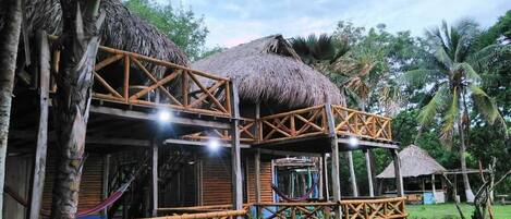A rustic thatched-roof structure with a bamboo railing, surrounded by lush greenery and palm trees.