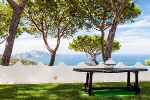 A table with a white vase and a metal lamp, set against a white wall with a view of the sea and mountains.