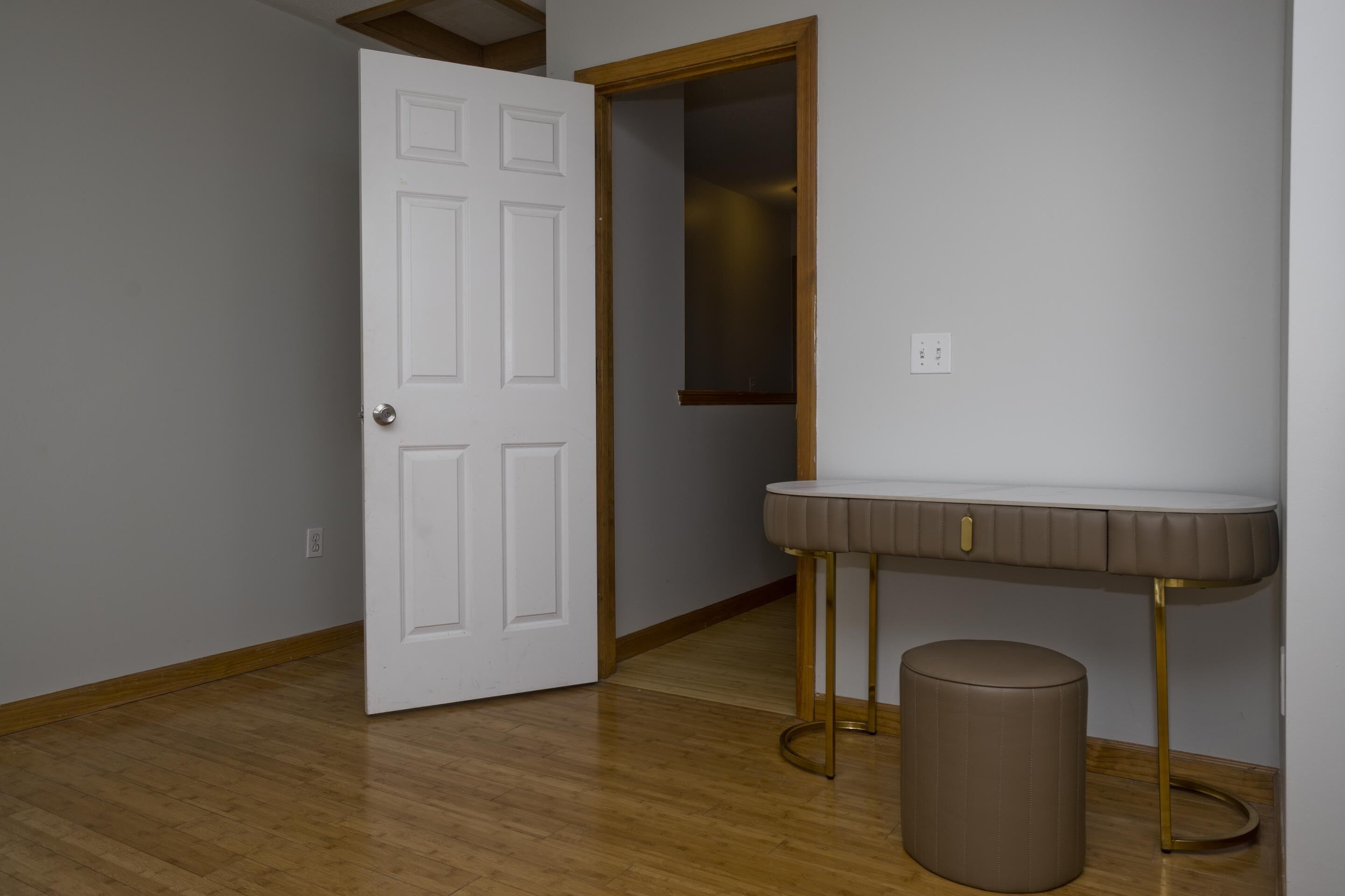 A room with a wooden floor, a white door, a vanity with a mirror, and a round stool.