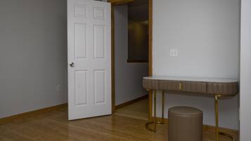 A room with a wooden floor, a white door, a vanity with a mirror, and a round stool.