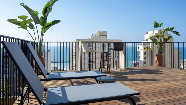 Une terrasse en bois avec des chaises longues et une table, d’où l’on a vue sur une étendue d’eau et des bâtiments.