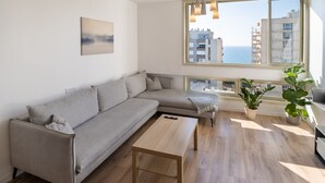 Un salon moderne avec un canapé d’angle gris, une table basse en bois et une grande fenêtre donnant sur la mer et les bâtiments.