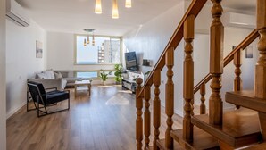 Un salon spacieux avec un escalier en bois, un ensemble de canapés, un téléviseur et une grande fenêtre donnant sur un paysage agréable.