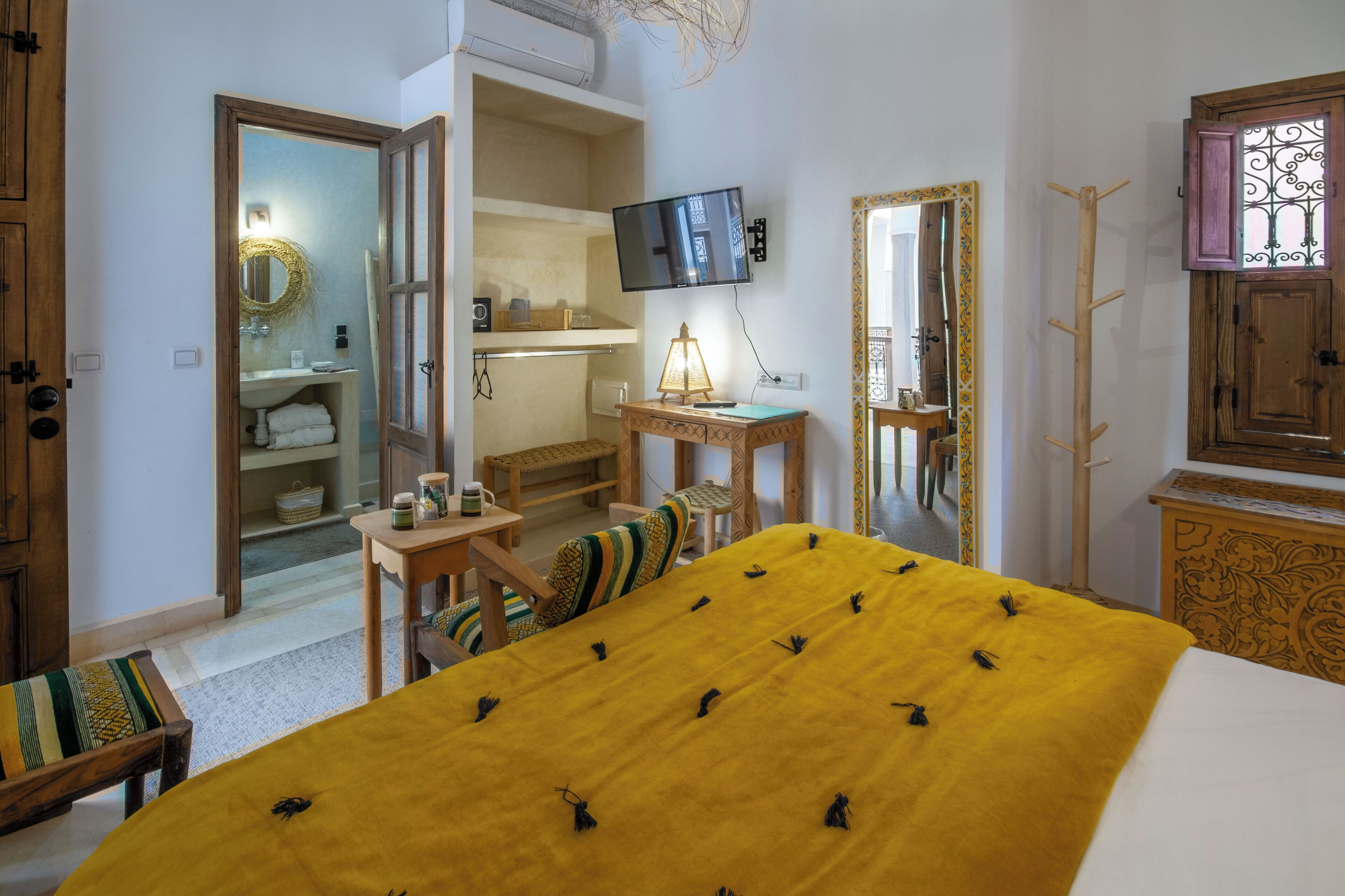 A hotel room with a bed, a television, a mirror, a wooden cabinet, and a small table with a lamp.
