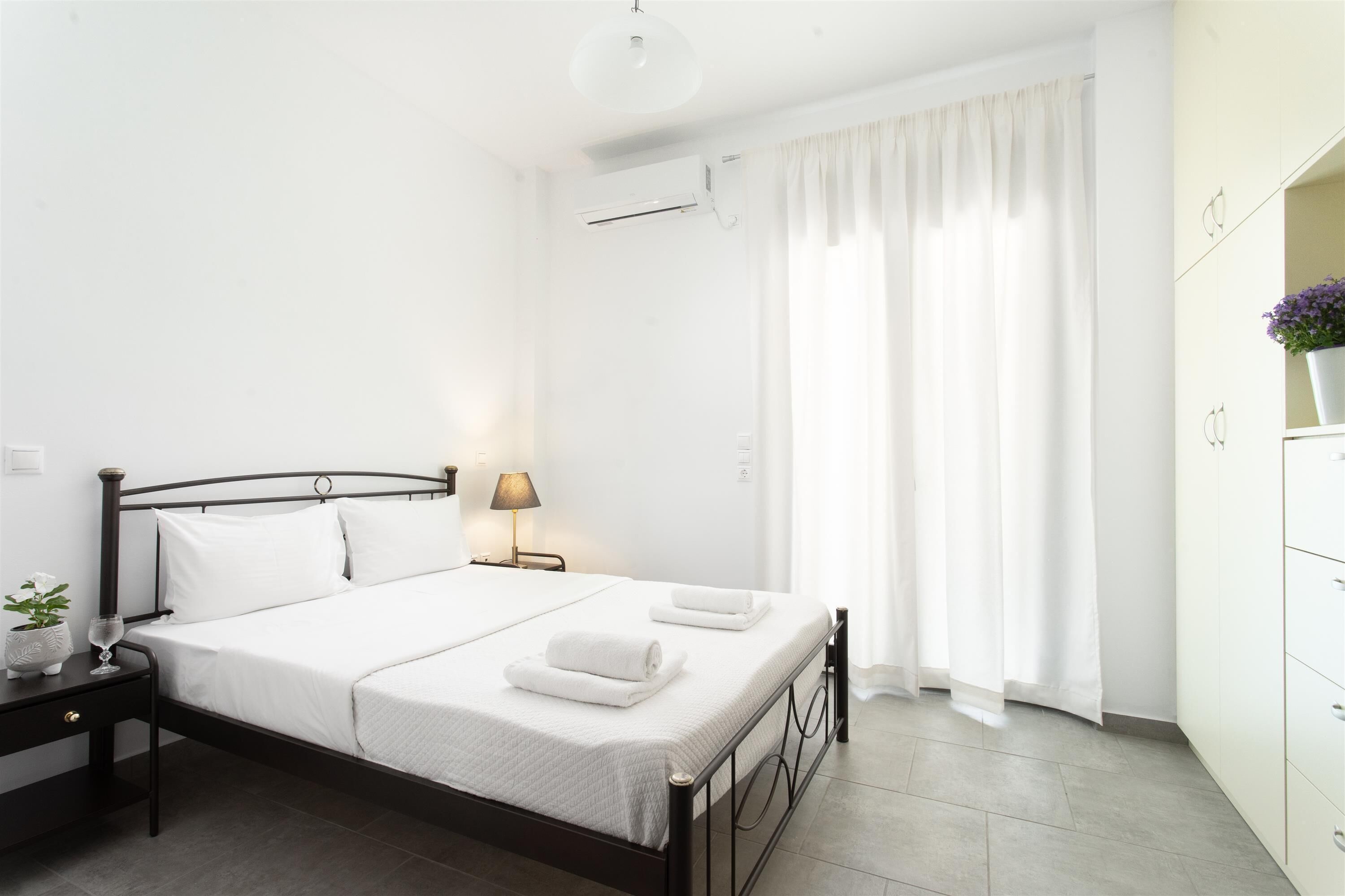 Un dormitorio ordenado con cama, mesita de noche, lámpara y ventana con cortinas.