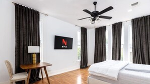 Un dormitorio con cama, escritorio, silla, ventilador de techo y un televisor que muestra el logotipo de Netflix.