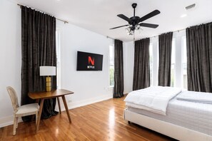 A bedroom with a bed, a desk, a chair, a ceiling fan, and a TV displaying the Netflix logo.