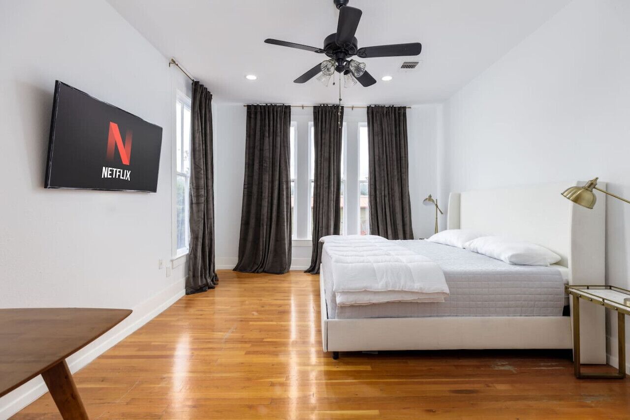 A bedroom with a bed, a ceiling fan, a TV mounted on the wall, and a window with curtains.