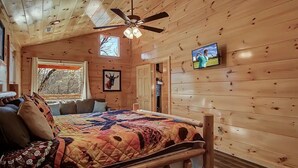 A cozy cabin room with a wooden ceiling, a bed with a colorful quilt, a ceiling fan, a TV on the wall, and a window with a view of trees. - Family Cabin w/ Game Room In Heart Of Pigeon Forge (Pigeon Forge)