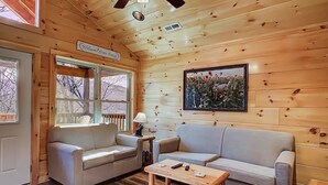 A cozy cabin interior with a wooden ceiling, a 'Welcome Home' sign, a framed picture of deer, a sofa, a coffee table, and a window with a view of trees. - Family Cabin w/ Game Room In Heart Of Pigeon Forge (Pigeon Forge)