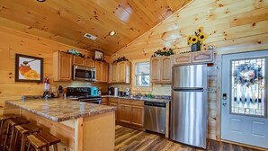 A wooden cabin interior with a kitchen featuring a granite countertop, stainless steel appliances, and wooden bar stools. - Family Cabin w/ Game Room In Heart Of Pigeon Forge (Pigeon Forge)