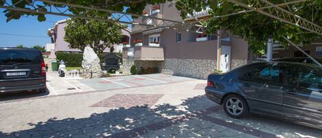 A street view with parked cars, a stone pathway, and a building with a stone facade and a balcony with potted plants.