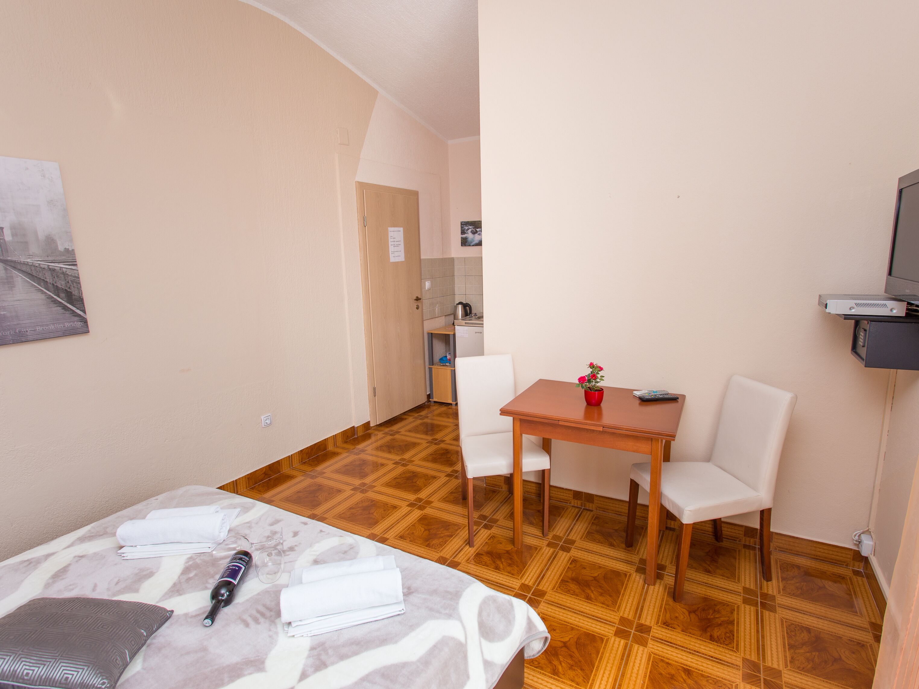 Habitación de hotel con cama, una mesita con un florero, televisión y una kitchenette al fondo.