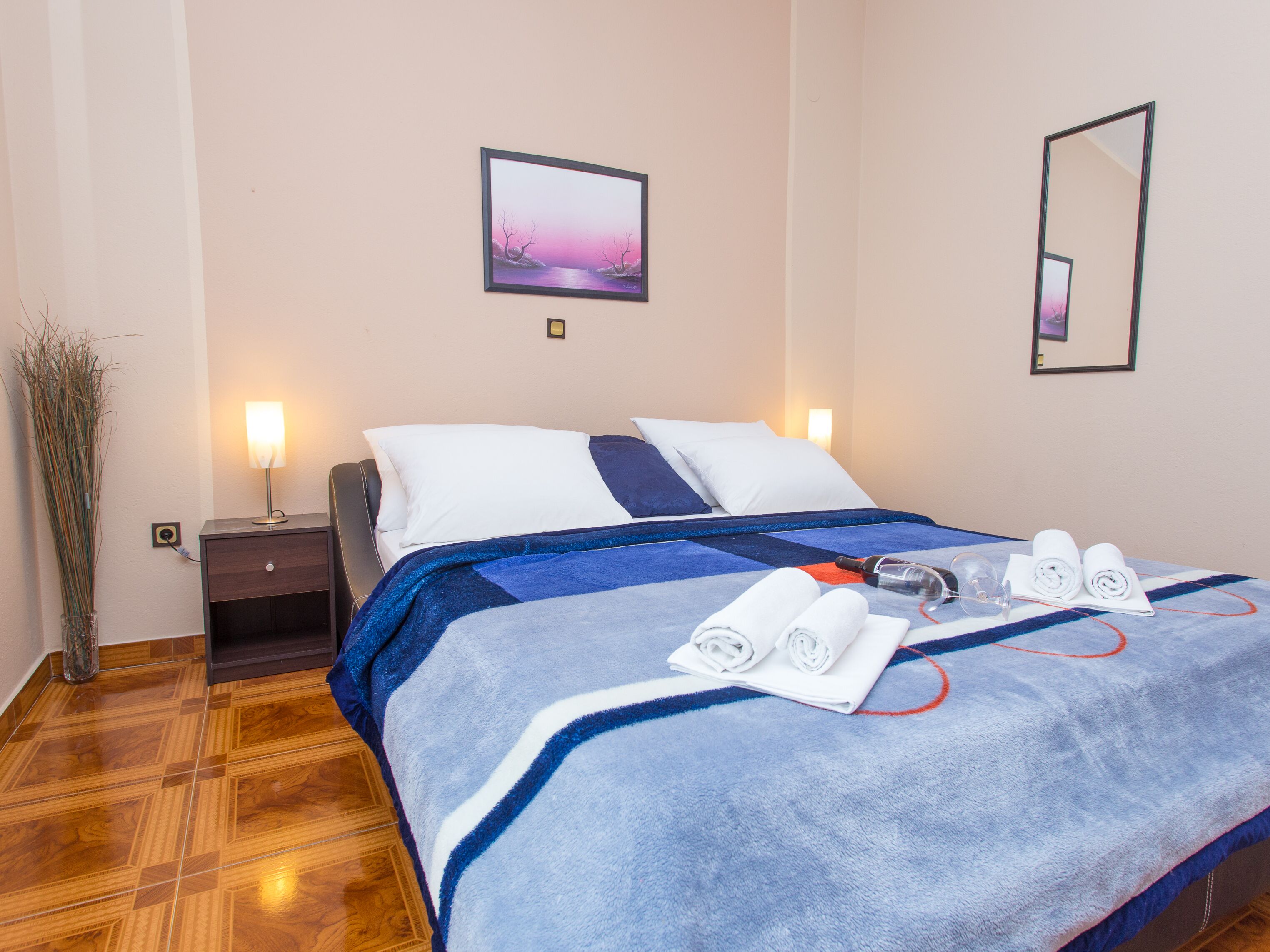 A neatly made bed with a blue and white blanket, two white pillows, and rolled towels on the bed. There is a nightstand with a lamp and a vase of dried plants beside the bed.
