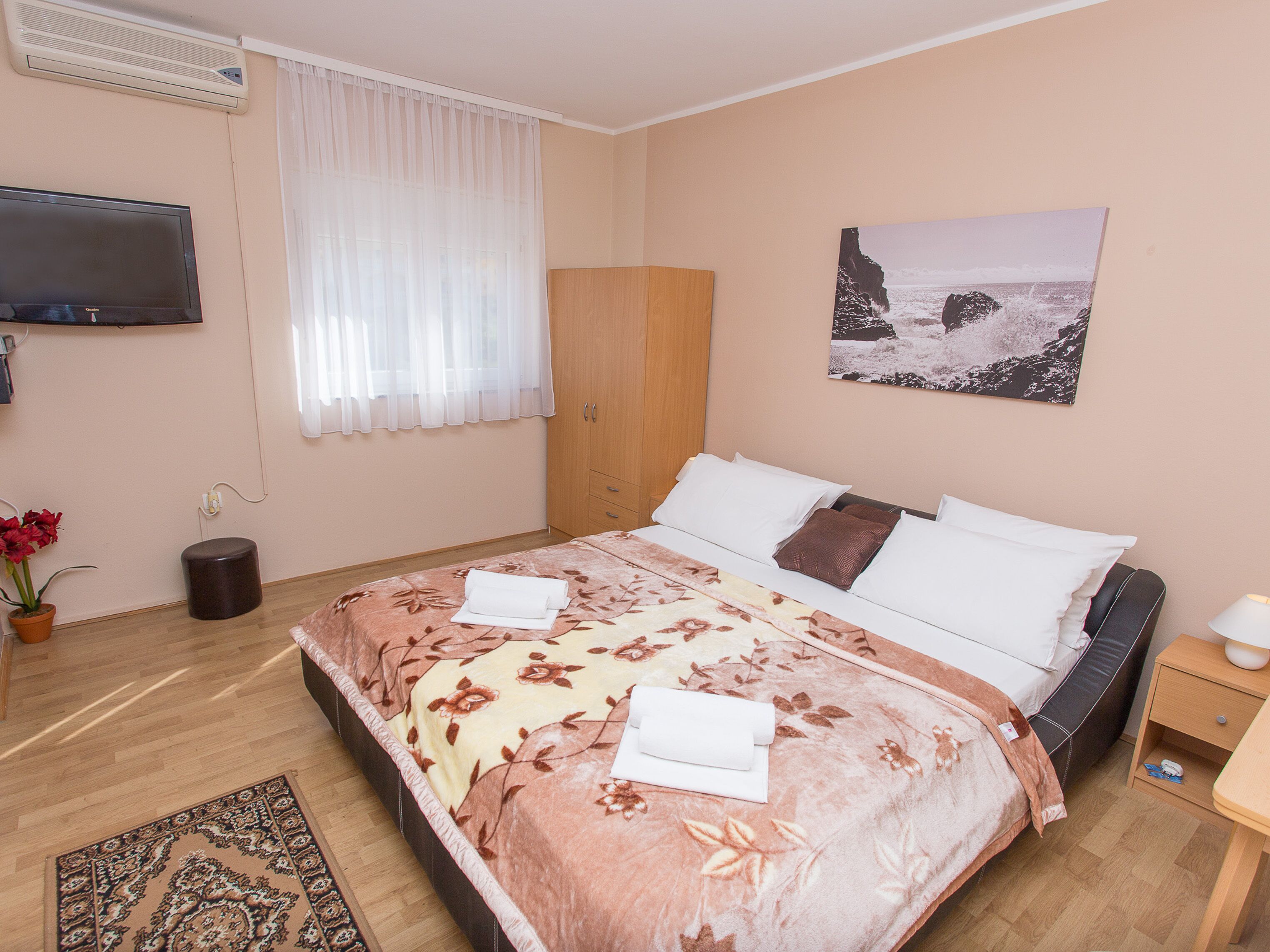 Une chambre à coucher comprenant un lit, un téléviseur, une armoire, un tableau, une plante en pot et un tapis.