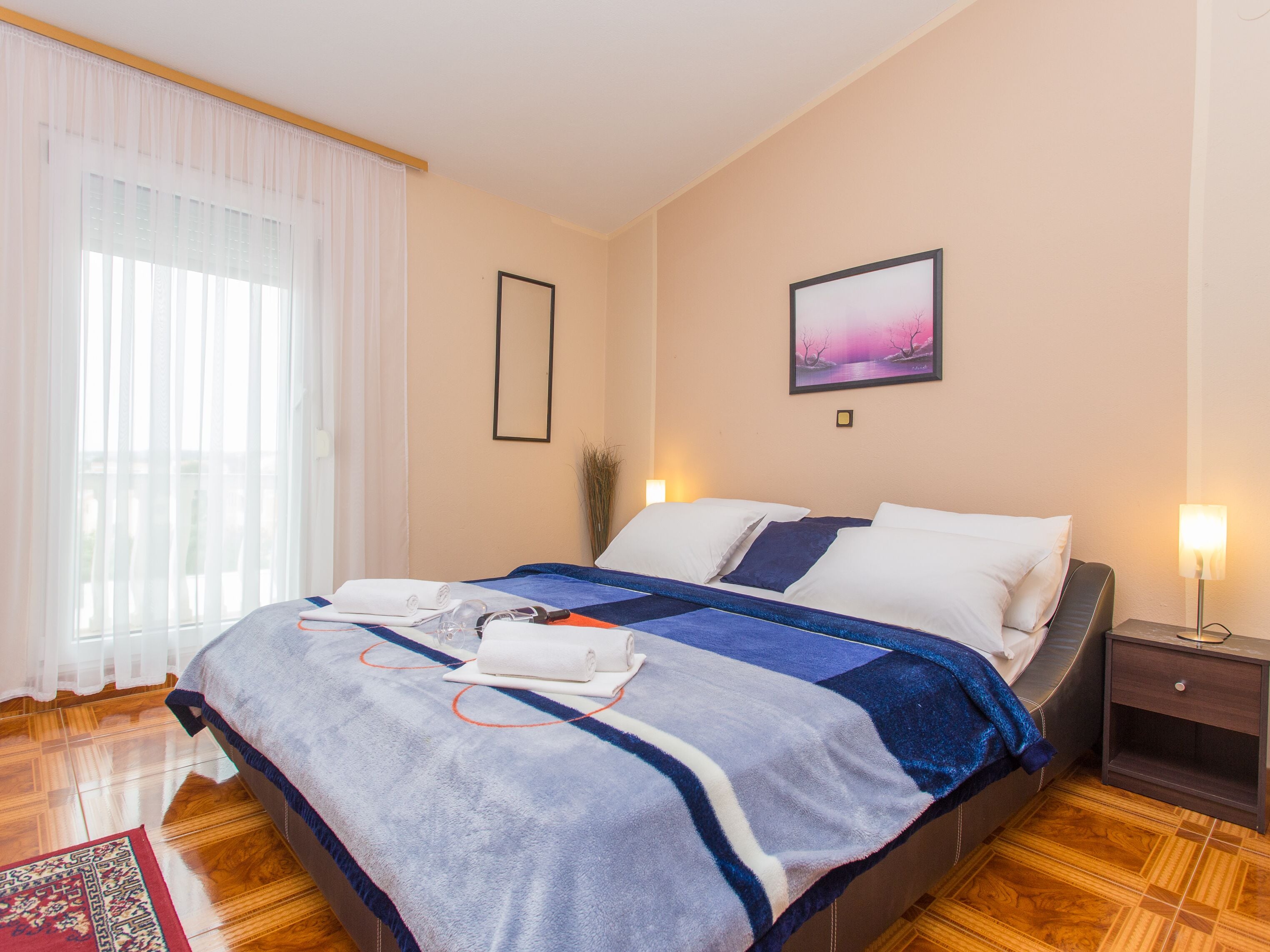 A hotel room with a bed, a nightstand, a lamp, a framed picture on the wall, and a window with curtains.