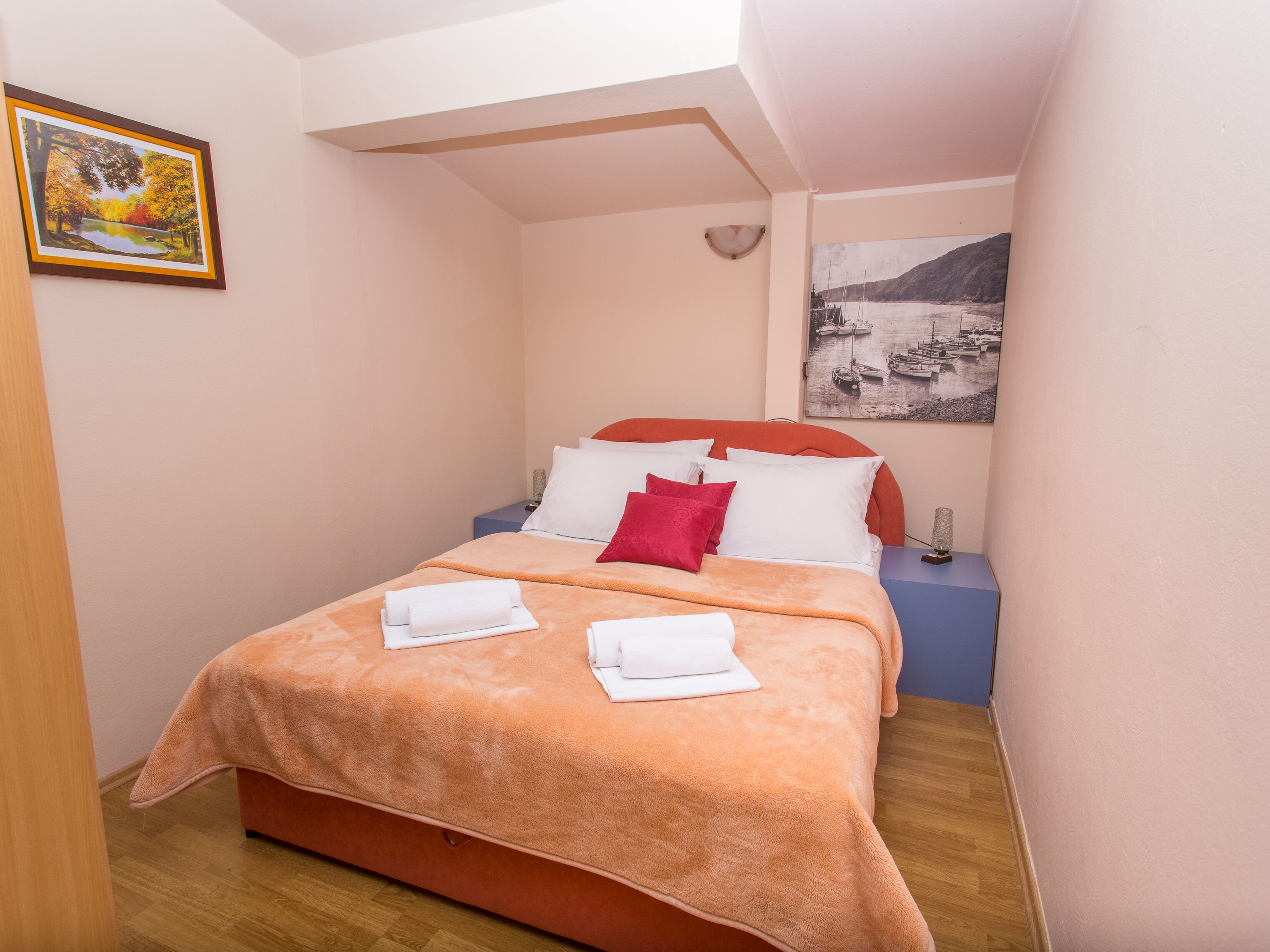 A neatly made bed with a peach-colored blanket, white pillows, and a red accent pillow. There is a framed picture of a landscape on the wall to the left and a black-and-white photo of boats on the right.