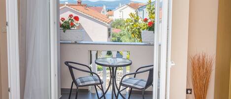 A room with a balcony, a bed, a table with chairs, and a view of buildings and mountains.