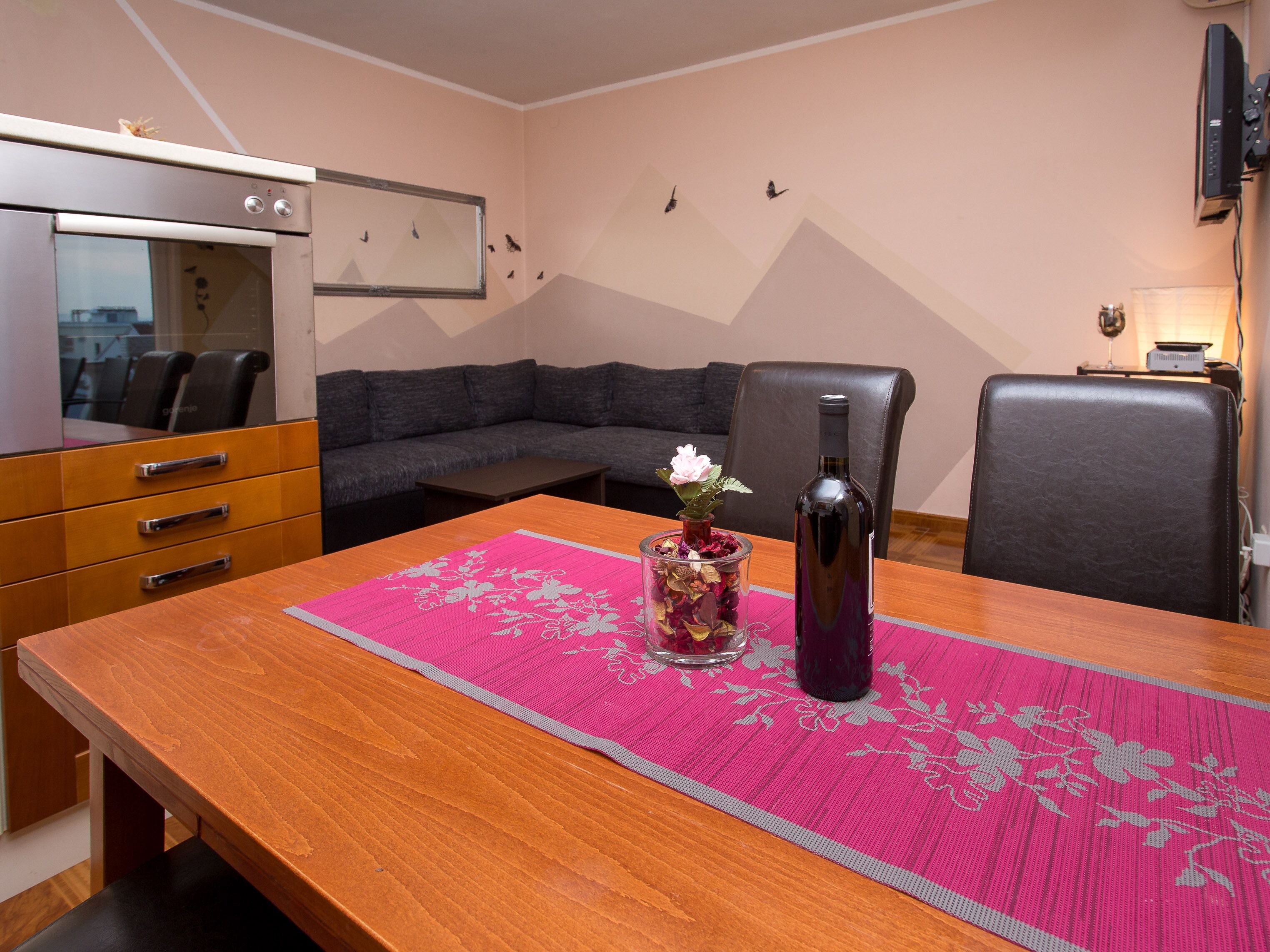 Un espacio habitable compacto con kitchenette, un sofá, una mesa de comedor con un mantel individual rosa y una botella de vino.