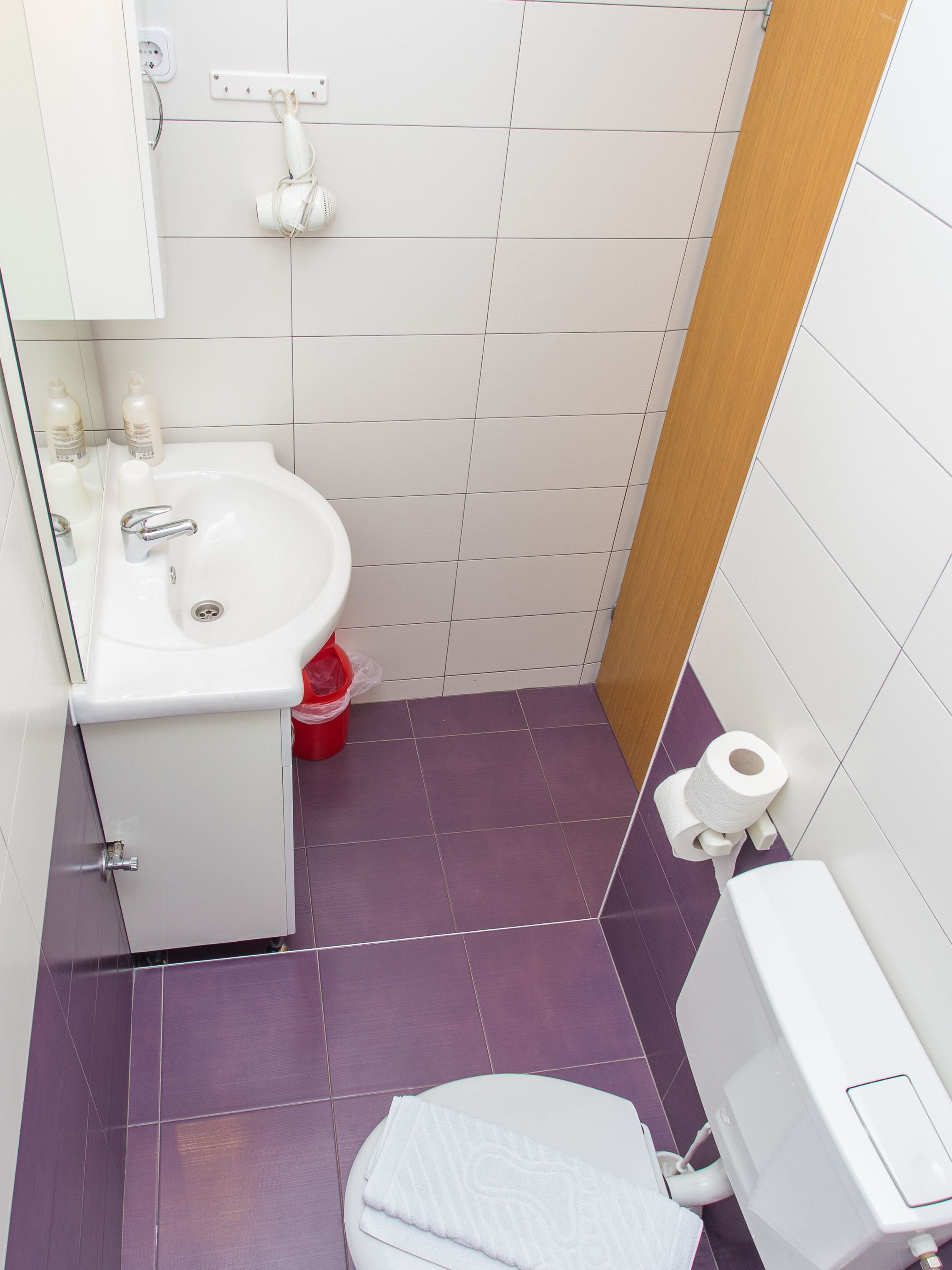 A small bathroom with a white sink, a toilet, and a urinal. There is a red bucket and a white towel on the floor.