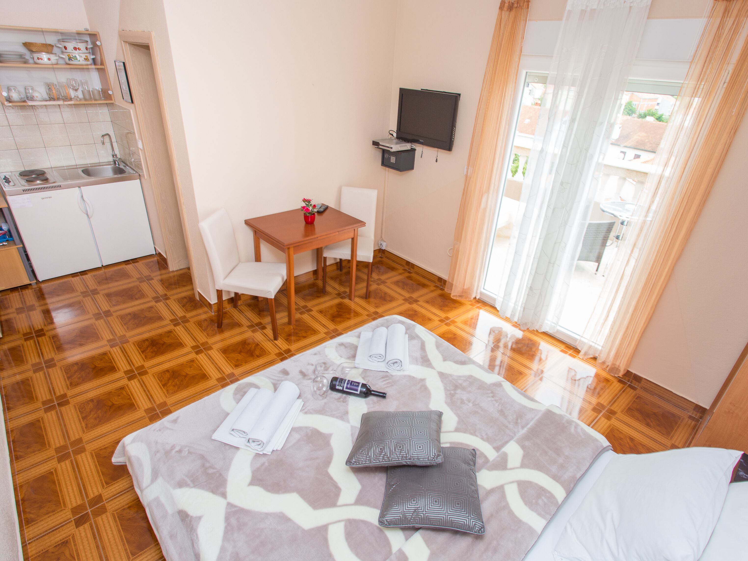A hotel room with a bed, a desk, a chair, a TV, a kitchen area, and a balcony with curtains.