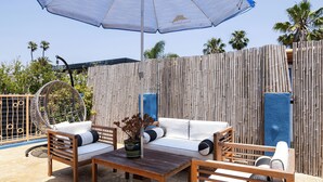 Un área de estar al aire libre con una mesa y sillas de madera, un sillón colgante y un paraguas azul.