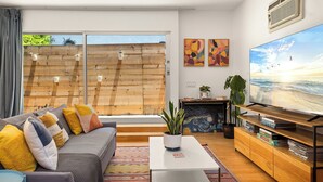 Una sala de estar moderna con un televisor grande, un sofá con cojines coloridos, una mesa de centro con una planta y un mueble de televisión de madera con juegos de mesa.