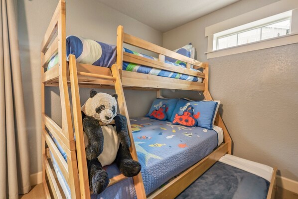 Una cama litera con un peluche de panda en la cama inferior, una colcha azul con peces y criaturas marinas, y una ventana sobre la cama.