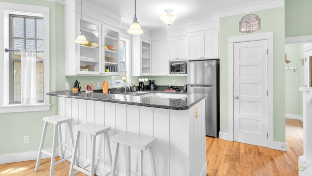 Una cocina moderna con armarios blancos, un refrigerador de acero inoxidable y una encimera de granito. Hay dos lámparas colgantes, un microondas y una cafetera en la encimera.