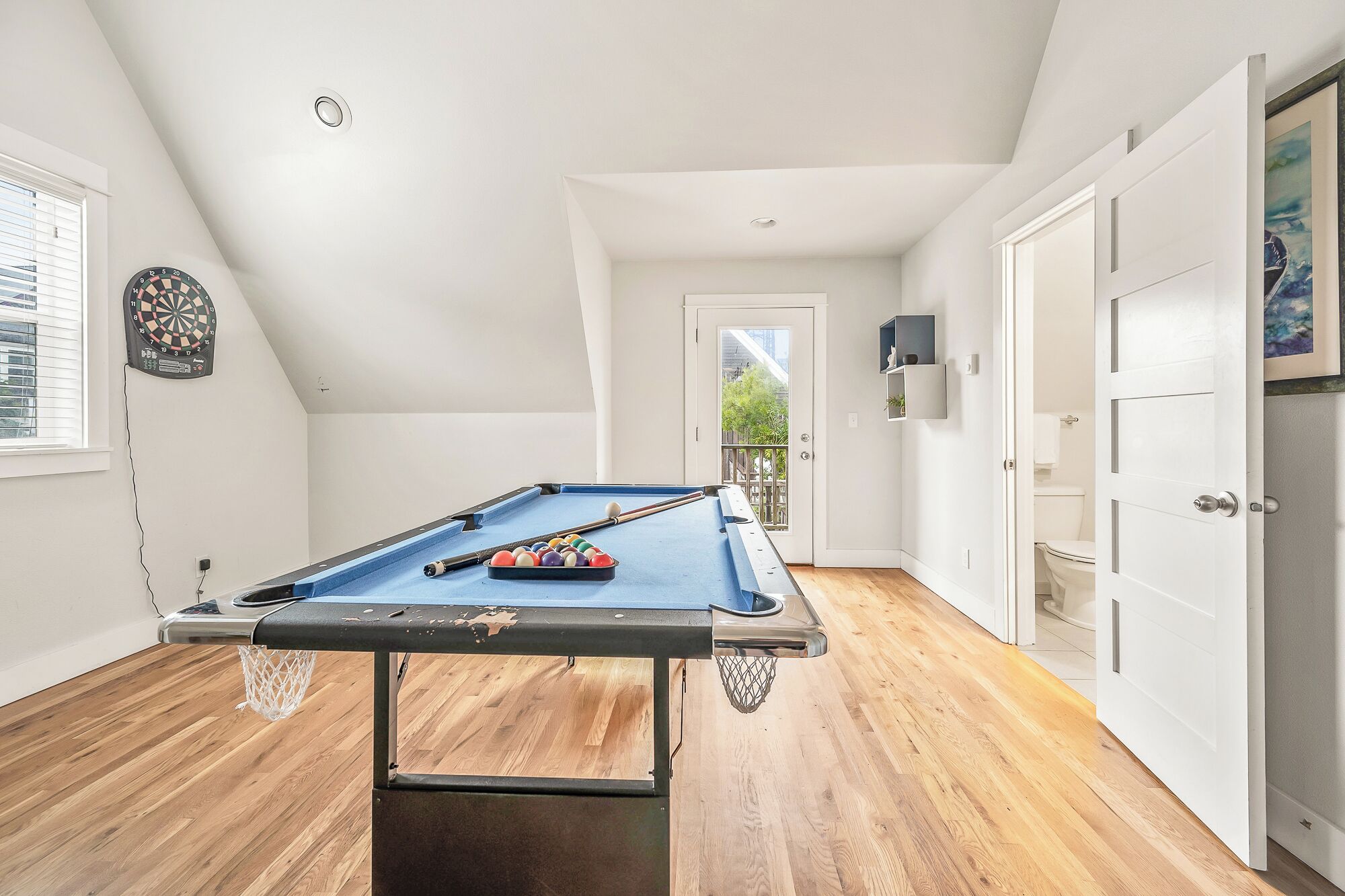 A pool table in a room with wooden flooring, a dartboard, and a door leading to a balcony.