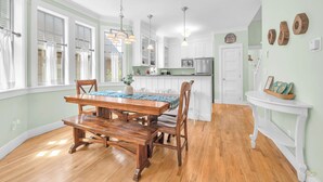 Un comedor amplio con una mesa y sillas de madera, una cocina con armarios blancos, y vistas al exterior a través de amplios ventanales.