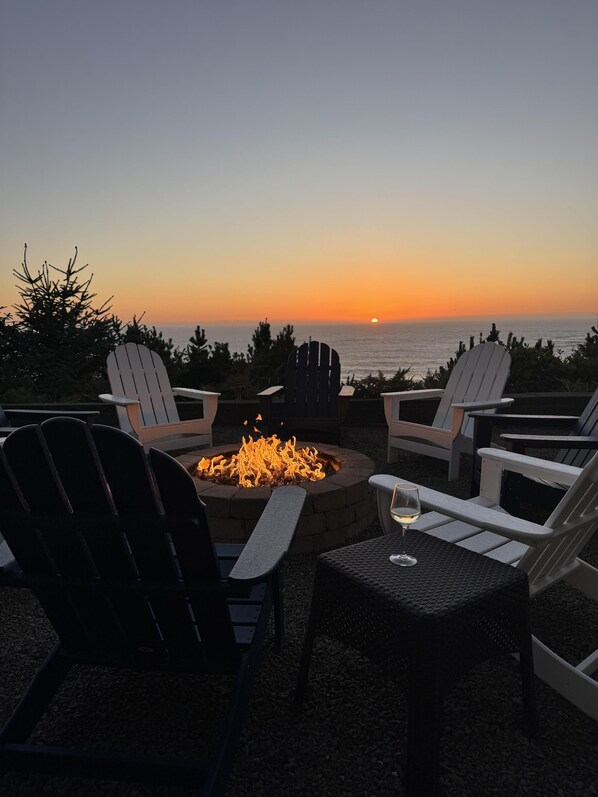 Un pozo de fuego rodeado por cuatro sillas Adirondack, con vista a la puesta de sol sobre el océano.