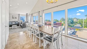 A dining area with a wooden table and white chairs, large windows with a view of greenery and a parking lot, and a chandelier. - The Flip Flop Inn - Walk to the Beach (Lincoln City)