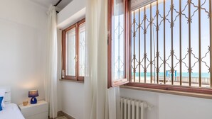 Een slaapkamer met een bed, nachtkastje, lamp, raam met uitzicht op het strand en een radiator.