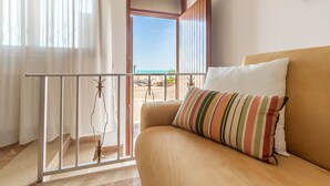 Een kamer met een beige bank, een gestreept kussen, uitzicht op het strand en een balkon met een leuning.