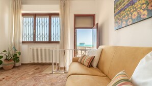Een woonkamer met een beige bank, een potplant, een radiator en uitzicht op het strand door een raam.