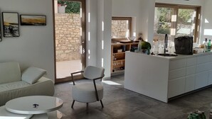 A modern living room with a sofa, a round coffee table, and a white chair. There is a kitchen area with a counter, a sink, and various kitchen items. A stone wall and a door leading outside are visible. - Exclusive Holiday Home Villa Marumare (Hvar)