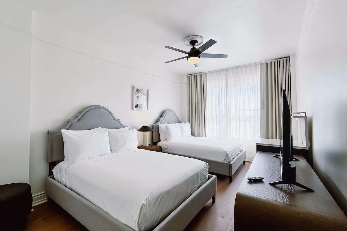 Ein Hotelzimmer mit zwei Betten, einem Deckenventilator, einem Nachttisch mit Lampe, einem Fernsehschrank und einem Fenster mit Vorhängen.