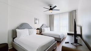 A hotel room with two beds, a ceiling fan, a bedside table with a lamp, a TV stand, and a window with curtains. - FOUND Hotel Carlton, Nob Hill (San Francisco)