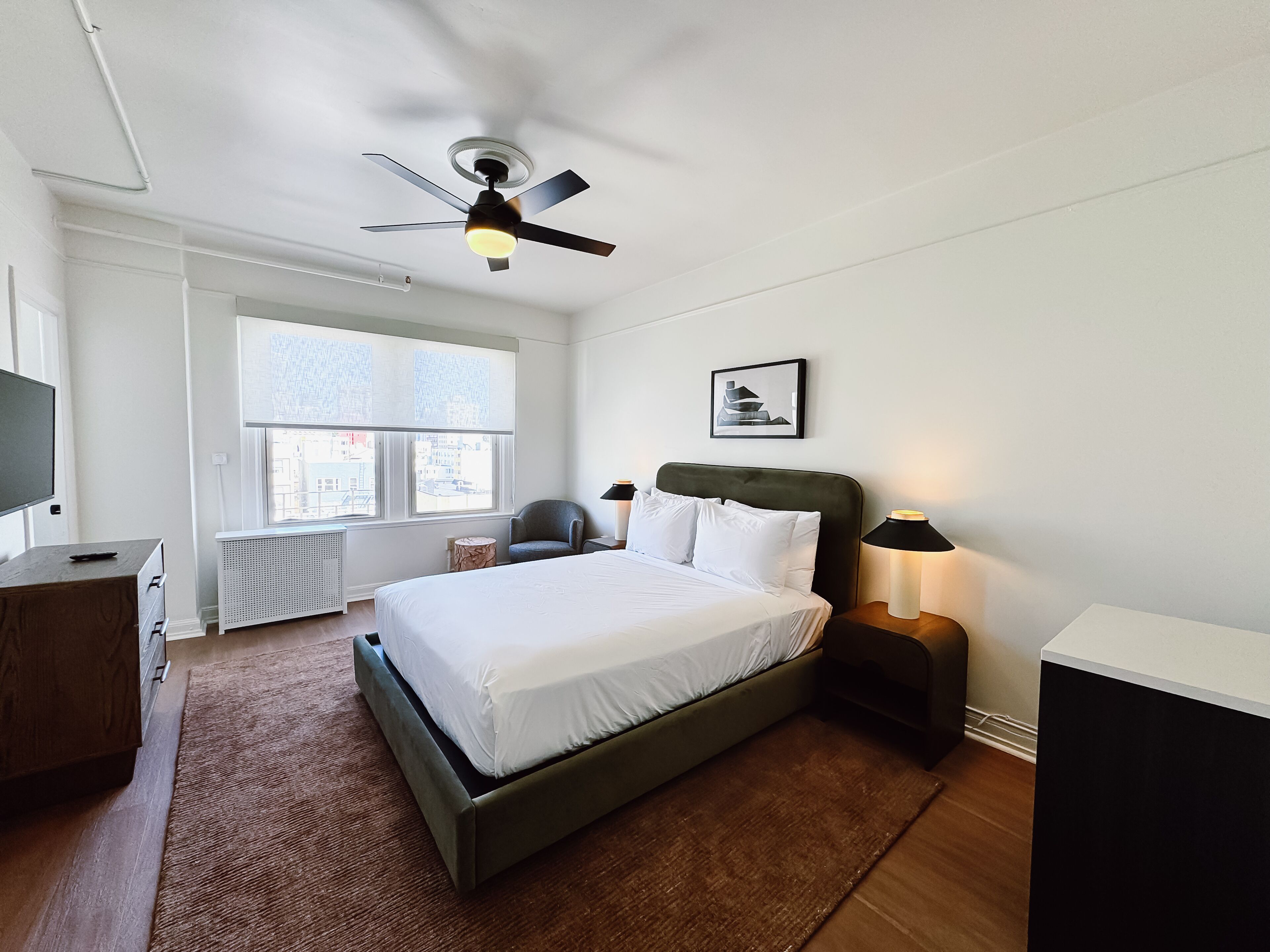 Une chambre d’hôtel comprenant un lit, un bureau, une chaise, un ventilateur de plafond, une fenêtre et une table de chevet avec une lampe.
