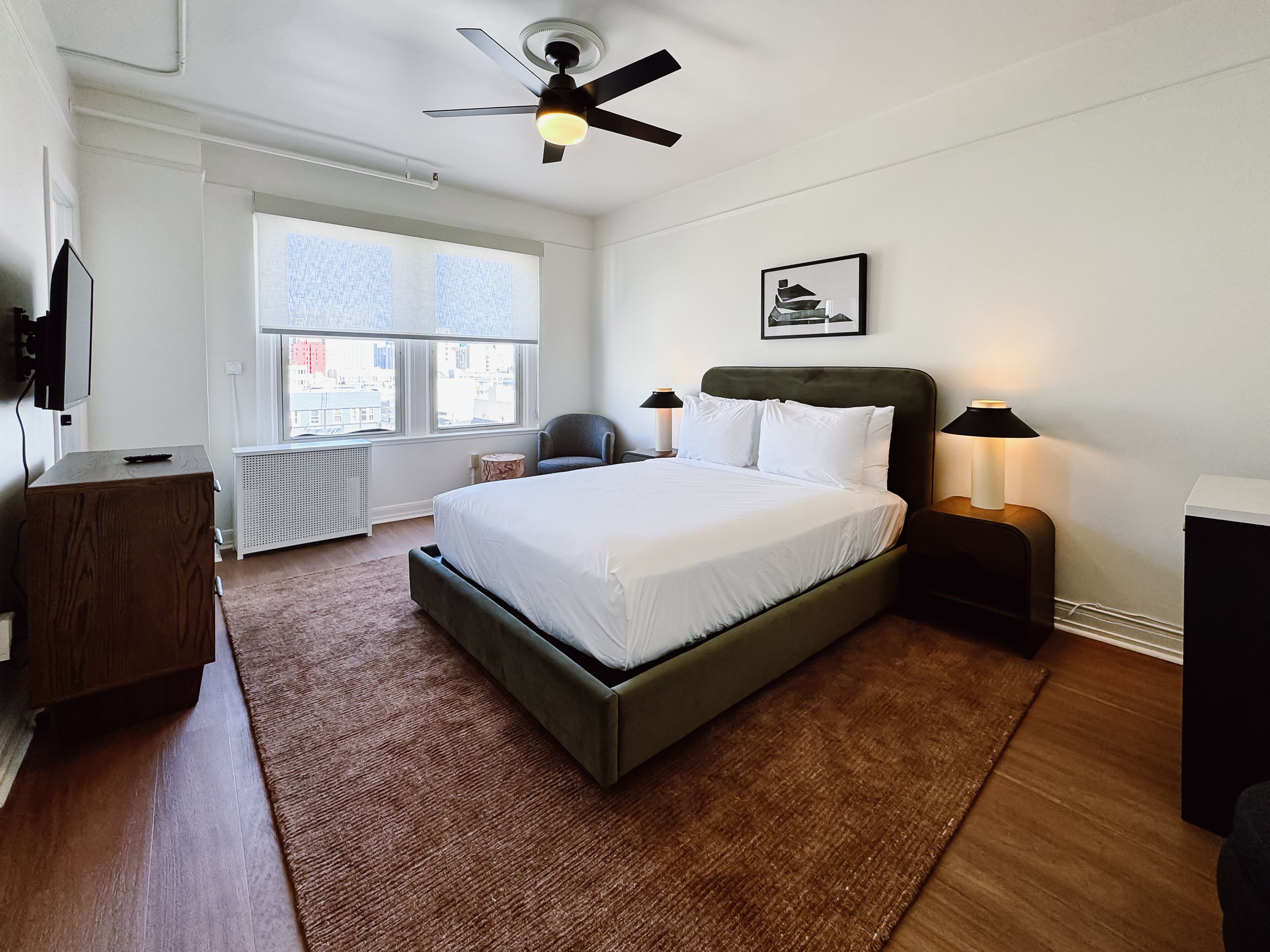 A modern bedroom with a large bed, a ceiling fan, a window with a city view, a chair, and bedside lamps.