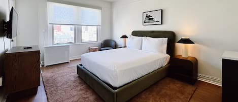 A modern bedroom with a large bed, a ceiling fan, a window with a city view, a chair, and bedside lamps.