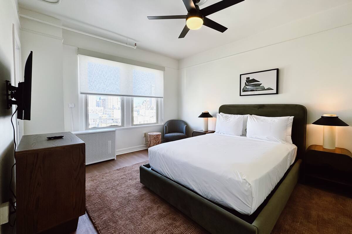 Ein Hotelzimmer mit Bett, Schreibtisch, Stuhl, Deckenventilator und einem Fenster mit Blick auf die Stadt.
