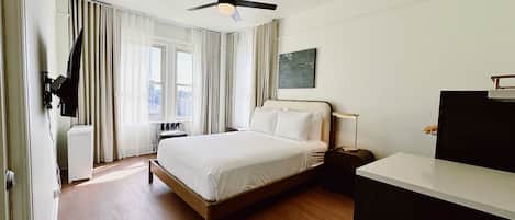 A modern hotel room with a large bed, a ceiling fan, a desk with a lamp, and a view of the city through the windows.