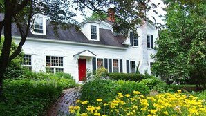 A white house with a red door, surrounded by lush greenery and yellow flowers. - Admiral Peary Inn (Fryeburg)